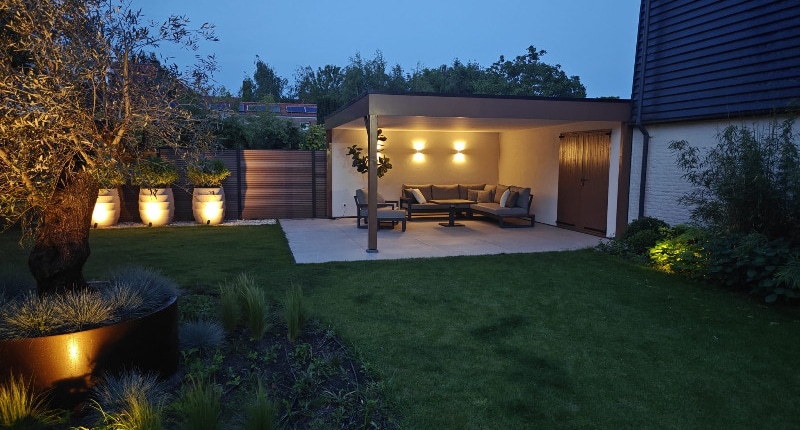 Overkapping-tuinrenovatie-waalwijk-van-hout Avondsfeer overkapping Waalwijk met up-down verlichting, loungebank en verlichte Luca Lifestyle potten